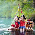 Kids dressed in pirate costumes and hats with treasure chest, spyglasses, and swords playing on wooden raft sailing in a river on hot summer day. Pirates role game for children. Water fun for family.